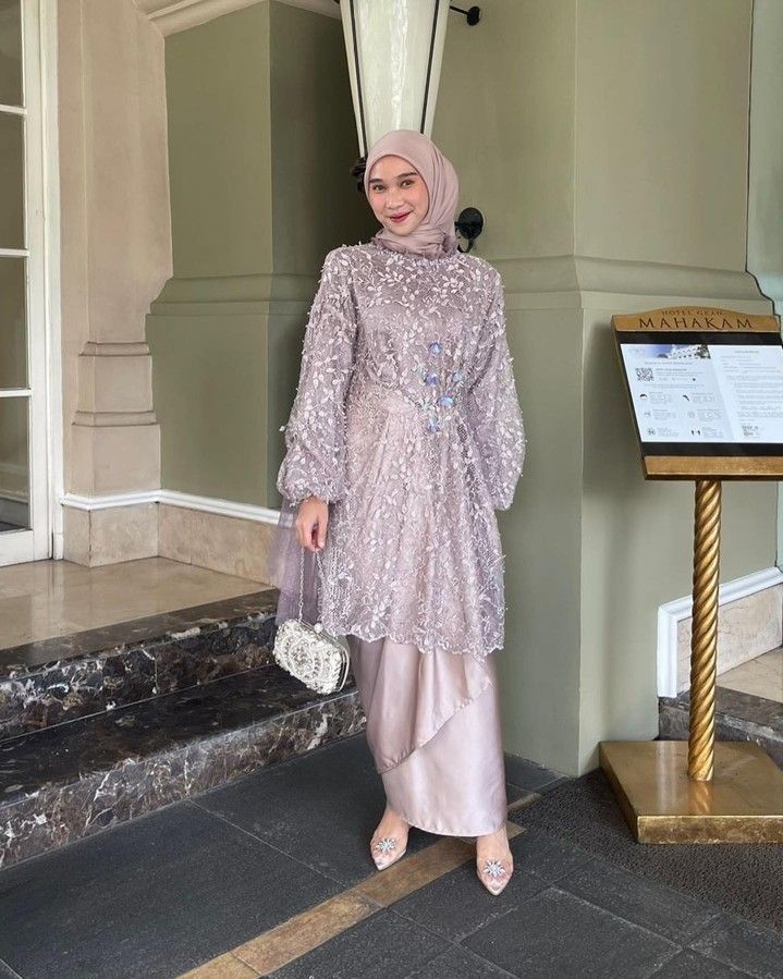 Wanita memakai kebaya brokat modern lengan panjang pastel ungu muda dengan hijab, membawa tas kecil
